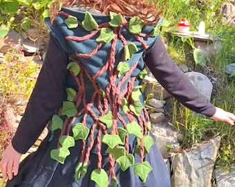 May include: A dark teal vest with a brown vine design and green leaf accents. The vest is worn over a black long-sleeved top and a dark skirt. The design resembles a tree, with the vines and leaves creating a nature-inspired look.