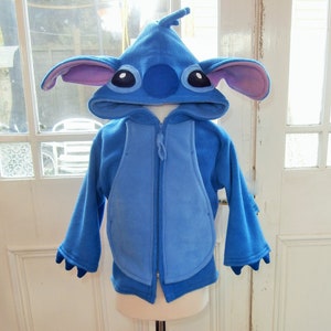 May include: A blue fleece hoodie with a character design, featuring large ears with pink inner details, large eyes, and a small antenna. The jacket has a zip closure and a light blue panel on the front. The sleeves end in claw-like details.