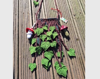 May include: A handmade brown shoulder bag with green leaf and mushroom accents. The bag features a woven design with hanging green leaves and decorative mushroom charms. The bag is made of fabric and has a long strap.