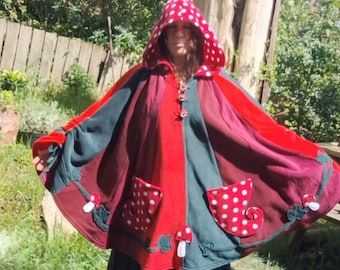 May include: A red, green, and burgundy hooded cloak with white polka dots and mushroom appliqués. The cloak has pockets with white polka dots and is worn outdoors. Text on the image reads "POV: Your portal to fairyland is open."