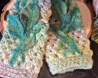 May include: Handmade fingerless gloves in a crochet style. The gloves are primarily light green, with accents of pink, blue, and orange. The design includes green leaf appliqués. The gloves are displayed on a wooden slice, with plants in the background.