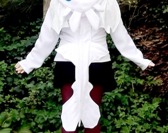 May include: White hooded jacket with blue eye details, resembling a cartoon character. The jacket has long sleeves and a zipper closure. The person is wearing a black skirt, burgundy leggings, and brown boots. The background is green foliage.