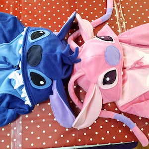 Puede incluir: Dos pijamas de una pieza de felpa, uno azul y otro rosa, con caras de personajes en las capuchas. El pijama azul tiene un dise&ntilde;o de Stitch, y el rosa, un dise&ntilde;o de Angel. Ambos tienen cremalleras.