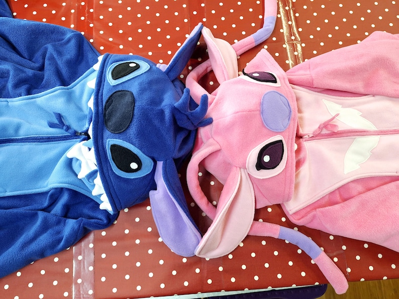 Puede incluir: Dos pijamas de una pieza de felpa, uno azul y otro rosa, con caras de personajes en las capuchas. El pijama azul tiene un dise&ntilde;o de Stitch, y el rosa, un dise&ntilde;o de Angel. Ambos tienen cremalleras.