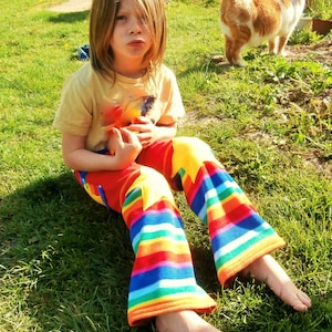 Puede incluir: Una persona joven con una camisa amarilla y pantalones a rayas arcoíris se sienta en un área cubierta de hierba. La persona está mirando a la cámara.