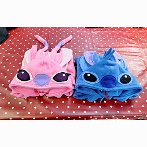 May include: Two character-themed hooded onesie pajamas, one pink and one blue. The pink onesie has a purple nose and ears, while the blue one has a blue nose. These cosy pajamas feature large character eyes and ears, perfect for lounging.
