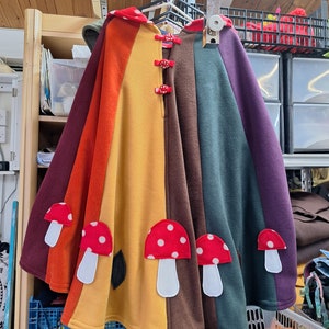 May include: A rainbow-colored fleece cape with a hood and mushroom appliques. The cape has red buttons and is hanging on a rack.