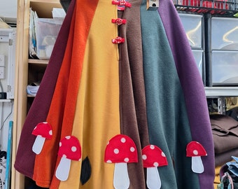 May include: A rainbow-colored fleece cape with a hood and mushroom appliques. The cape has red buttons and is hanging on a rack.