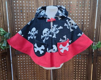 May include: A black and red hooded cape with a skull and crossbones pattern. The cape features two pockets with white crossbones and a red bow closure. The cape is designed for children.