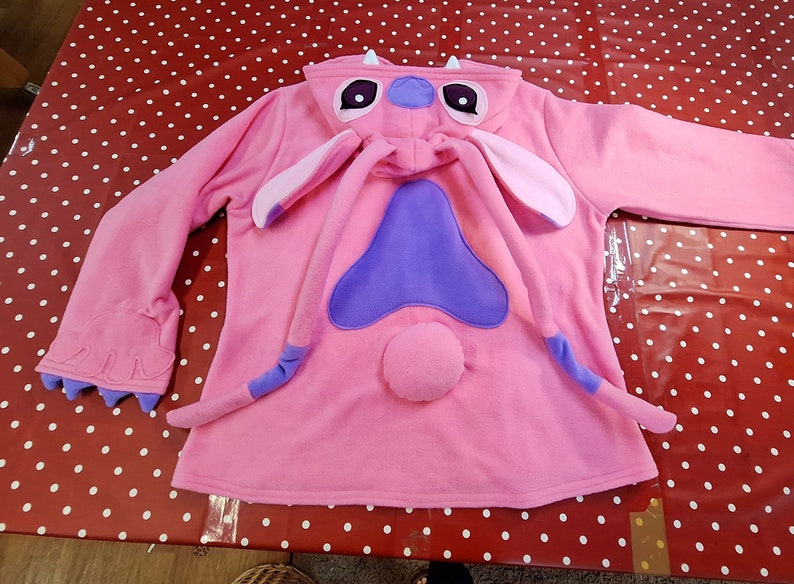 Puede incluir: Mono con capucha rosa con un dise&ntilde;o de personaje, con orejas grandes, una nariz morada y una cola. El mono tiene una capucha con ojos y antenas de personaje. Las mangas terminan en detalles de garras. Un atuendo divertido y tem&aacute;tico.