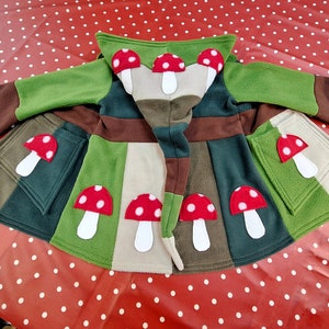 May include: A green, brown, and beige fleece jacket with a hood and mushroom appliques. The jacket has pockets on the sides and a drawstring at the waist.