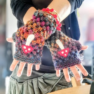 Può includere: Un paio di guanti senza dita all'uncinetto colorati con applicazioni a forma di fungo. I guanti sono realizzati con una varietà di colori, tra cui rosso, arancione, giallo, verde, blu e viola. I funghi sono bianchi con cappelli rossi e punti bianchi.