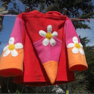 Peut inclure: Une veste en polaire rouge avec un motif de marguerite rose, orange et blanche. La veste a une capuche et des manches longues.