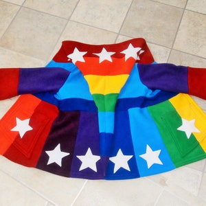 Puede incluir: Una colorida capa con temática de arcoíris con detalles de estrellas blancas. La capa presenta un vibrante espectro de colores, incluyendo rojo, naranja, amarillo, verde, azul y morado. Estrellas blancas están cosidas en la capa. La capa tiene mangas y bolsillos.