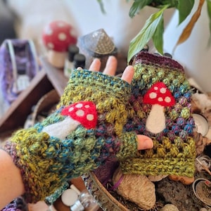 Puede incluir: Un par de guantes sin dedos hechos a mano en un patrón de ganchillo texturizado y colorido. Cada guante presenta una aplicación de hongo rojo con puntos blancos y un tallo blanco. Los guantes tienen una mezcla de hilo verde, azul y amarillo.