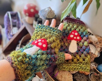 May include: A pair of handmade fingerless gloves in a colorful, textured crochet pattern. Each glove features a red mushroom applique with white spots and a white stem. The gloves have a mix of green, blue, and yellow yarn.