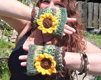 May include: A pair of handmade crochet wrist cuffs. Each cuff features a vibrant yellow sunflower with a brown center, set against a backdrop of green, brown, and cream-colored yarn. The cuffs are designed to be worn around the wrists.