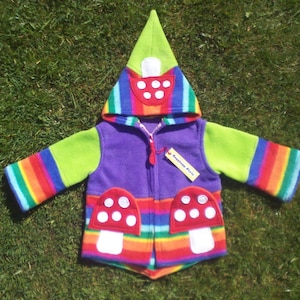 Puede incluir: Una chaqueta de forro polar morada con un patrón de rayas arcoíris y una capucha verde puntiaguda. La chaqueta tiene dos bolsillos en forma de hongo con tapas rojas y tallos blancos.