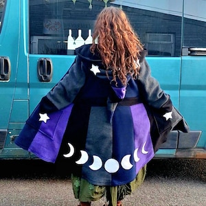 May include: A hooded cloak with a celestial design. The cloak features a color block design in shades of black, gray, and purple, with white stars and moon phases. The person is wearing green pants and brown boots.