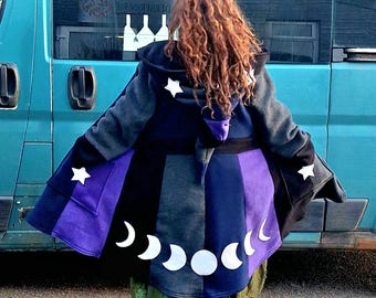 May include: A hooded cloak with a celestial design. The cloak features a color block design in shades of black, gray, and purple, with white stars and moon phases. The person is wearing green pants and brown boots.