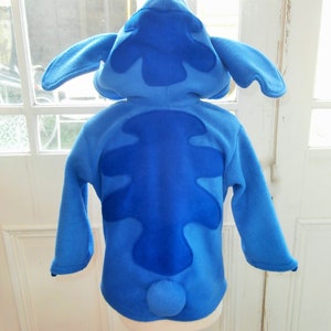 May include: A blue fleece costume, styled after the character Stitch. It has a hood with large ears, a dark blue patch on the back, and a small, round tail. The long-sleeved costume is displayed on a mannequin, against a window.