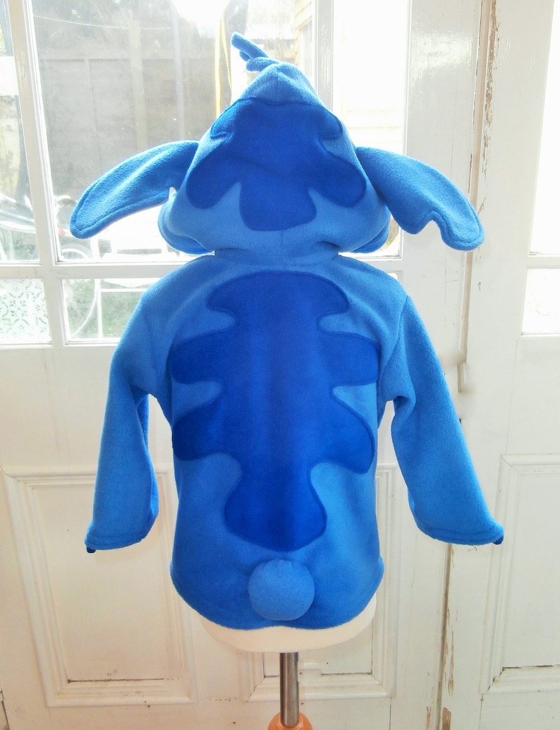 May include: A blue fleece costume, styled after the character Stitch. It has a hood with large ears, a dark blue patch on the back, and a small, round tail. The long-sleeved costume is displayed on a mannequin, against a window.