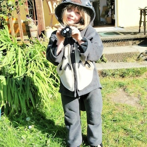 May include: A child wearing a gray Totoro-inspired costume, complete with a hooded jacket, pants, and a plush toy. The costume features a cream-colored belly and black accents. The child is standing on green grass.