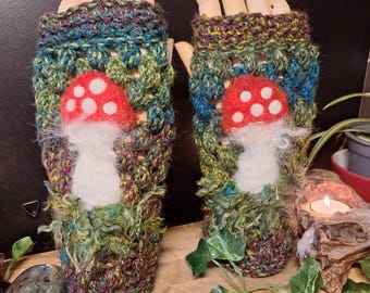 May include: A pair of handmade fingerless gloves. The gloves are crocheted with a mix of green, blue, and purple yarn. Each glove features a red mushroom with white spots. The gloves are displayed on wooden hand forms.