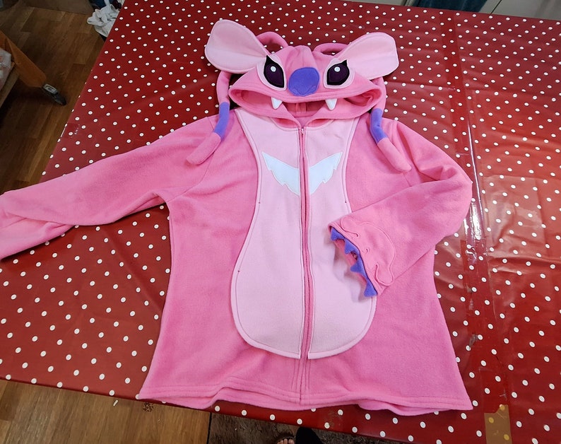 Puede incluir: Una sudadera con capucha rosa con cremallera que representa al personaje Angel de Lilo & Stitch. La sudadera tiene una capucha con orejas grandes, detalles morados y un dise&ntilde;o espec&iacute;fico del personaje en la parte delantera. Las mangas tienen un dise&ntilde;o de pu&ntilde;o &uacute;nico.