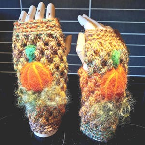 Peut inclure: Une paire de mitaines faites à la main. Les gants sont tricotés avec un fil texturé multicolore dans les tons de marron, d'orange et de vert. Chaque gant présente une citrouille orange feutrée avec une feuille verte. Les gants sont conçus pour l'automne ou Halloween.