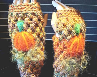 May include: A pair of handmade fingerless gloves. The gloves are knitted with a textured, multicolored yarn in shades of brown, orange, and green. Each glove features a felted orange pumpkin with a green leaf. The gloves are designed for fall or Halloween.