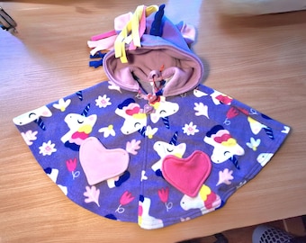 May include: A purple fleece unicorn cape with a hood, featuring unicorn graphics, pink hearts, and floral accents. The hood has a blue horn and colorful mane. A toggle closure secures the cape.