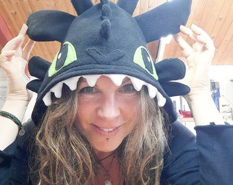 May include: A woman wearing a black fleece hooded hat with a dragon design. The hat has green eyes, white teeth, and black wings. The woman is smiling and has long brown hair.