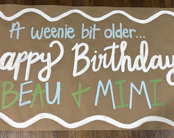 Puede incluir: Un letrero de papel marrón con letras blancas, azules y verdes. El letrero dice "A weenie bit older... happy Birthday BEAU + MIMI". Hay ilustraciones de un perrito caliente y un perro sosteniendo globos.