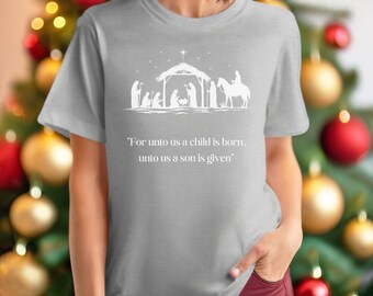 May include: A light gray t-shirt featuring a white graphic of a nativity scene with the text "For unto us a child is born, unto us a son is given." The shirt has short sleeves and a crew neck.