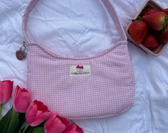 May include: A pink gingham shoulder bag with a matching handle and a small charm. The bag has a label that reads "o teddy bear totes". Fresh strawberries in a small pink dish and pink tulips are also in the image.