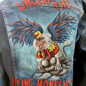 May include: A denim jacket with a vibrant, hand-painted design on the back. The artwork features a winged monkey with a fierce expression, set against a blue backdrop. The words "UNLEASH THE FLYING MONKEY!" are written in red.