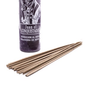 May include: A purple candle with a white image of a man and the text "Juan el Conquistador" and "Bendicion al Hogar Veladora de Nove" on the front. A bundle of brown incense sticks are in the foreground.