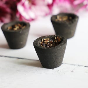 May include: Three dark gray, cone-shaped incense holders filled with dried herbs and spices. The holders are arranged on a white wooden surface, with pink flowers blurred in the background. The incense holders have a textured appearance.