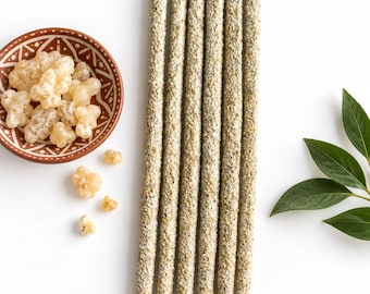 May include: Five long, cylindrical incense sticks with a textured, light beige and grey speckled coating, arranged vertically. To the left is a small, round, terracotta-coloured bowl with white geometric patterns containing pale resin chunks.