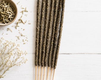 May include: Six dark green, textured incense sticks with smooth wooden bases arranged vertically. A small bowl containing dried green herbs and a sprig of dried white flowers are positioned to the left. The text ESOTERIC AROMA is visible.