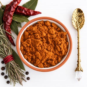 May include: A round ceramic bowl filled with vibrant orange ground spice, positioned next to a bundle of dried rosemary tied with red twine, several dried red chili peppers, green bay leaves, black peppercorns, and a decorative gold spoon.