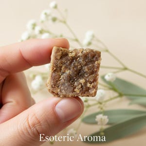 Hand holding a square piece amber resin incense with 'Esoteric Aroma' text, against a light background with green leaves.