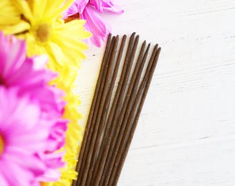 May include: A bundle of brown incense sticks arranged on a white wooden surface, with a vibrant display of yellow and pink flowers in the background. The incense sticks are slender and evenly spaced.