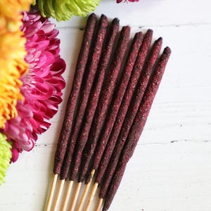 May include: A bundle of seven dark maroon incense sticks with wooden bases, arranged vertically next to a cluster of vibrant pink, orange, and lime green chrysanthemum flowers on a white surface.