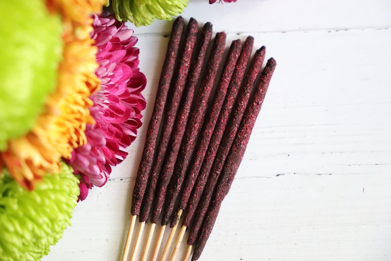 May include: A bundle of seven dark maroon incense sticks with wooden bases, arranged vertically next to a cluster of vibrant pink, orange, and lime green chrysanthemum flowers on a white surface.