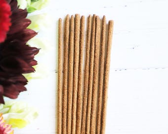 May include: A collection of brown incense sticks arranged vertically on a white wooden surface. The incense sticks are a light brown color and have a textured appearance. The image also features colorful flowers in the background.
