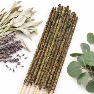 May include: A bundle of ten incense sticks with a textured, dark green and brown herbal coating, arranged vertically alongside sprigs of dried white sage, purple lavender flowers, and round green eucalyptus leaves on a white background.