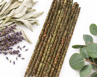 May include: A bundle of ten incense sticks with a textured, dark green and brown herbal coating, arranged vertically alongside sprigs of dried white sage, purple lavender flowers, and round green eucalyptus leaves on a white background.
