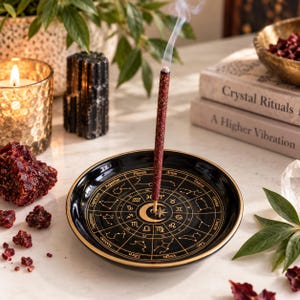 One lit Dragon's Blood mini incense stick releasing delicate smoke from a glossy black ceramic zodiac incense holder with gold celestial designs, surrounded by red resin, black tourmaline, greenery, and warm candlelight in an elegant home setting.
